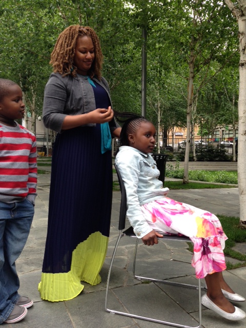 caption: Salamata Sylla demonstrates African hair braiding on her daughter while her son watches on.