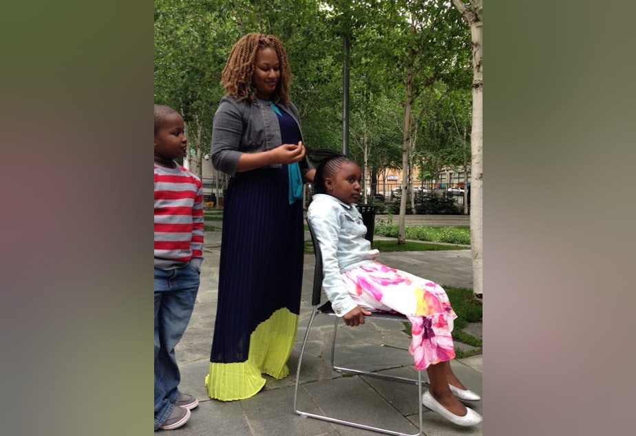 caption: Salamata Sylla demonstrates African hair braiding on her daughter while her son watches on.