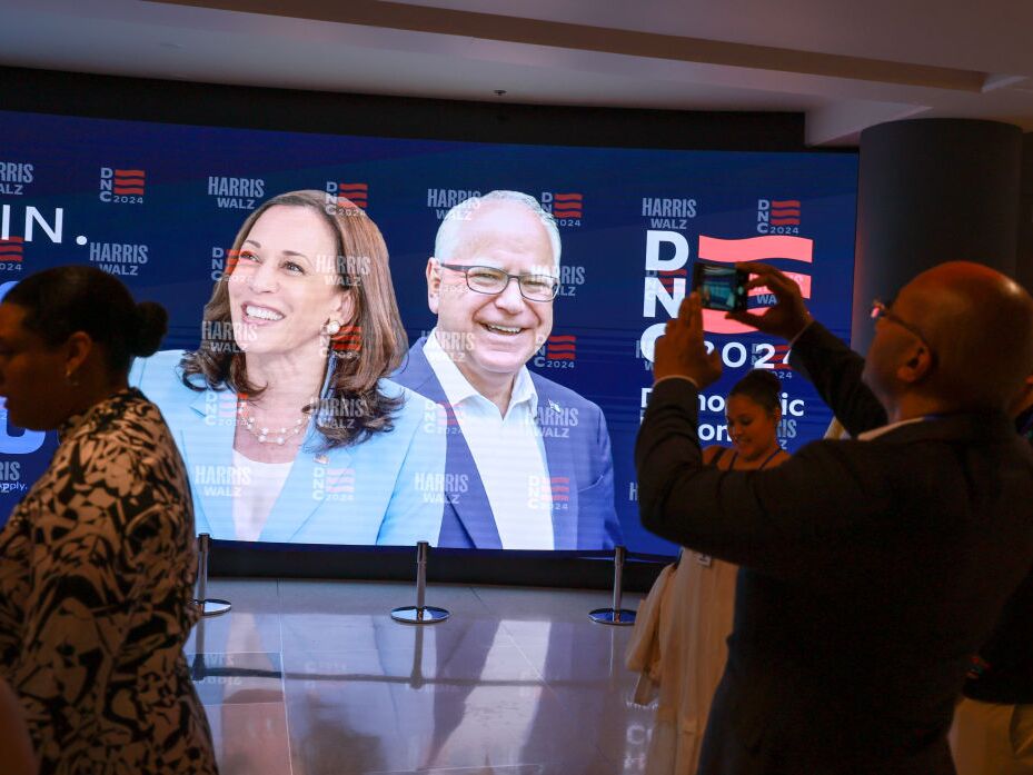 caption: People stand in front of a sign featuring Democratic presidential candidate Vice President Kamala Harris and Democratic vice presidential candidate Minnesota Gov. Tim Walz at the United Center before the start of the Democratic National Convention Friday in Chicago.