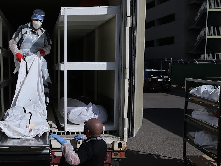 caption: A low-level inmate from El Paso County detention facility works loading bodies wrapped in plastic into a refrigerated temporary morgue trailer in a parking lot of the El Paso County Medical Examiner's office on Tuesday.