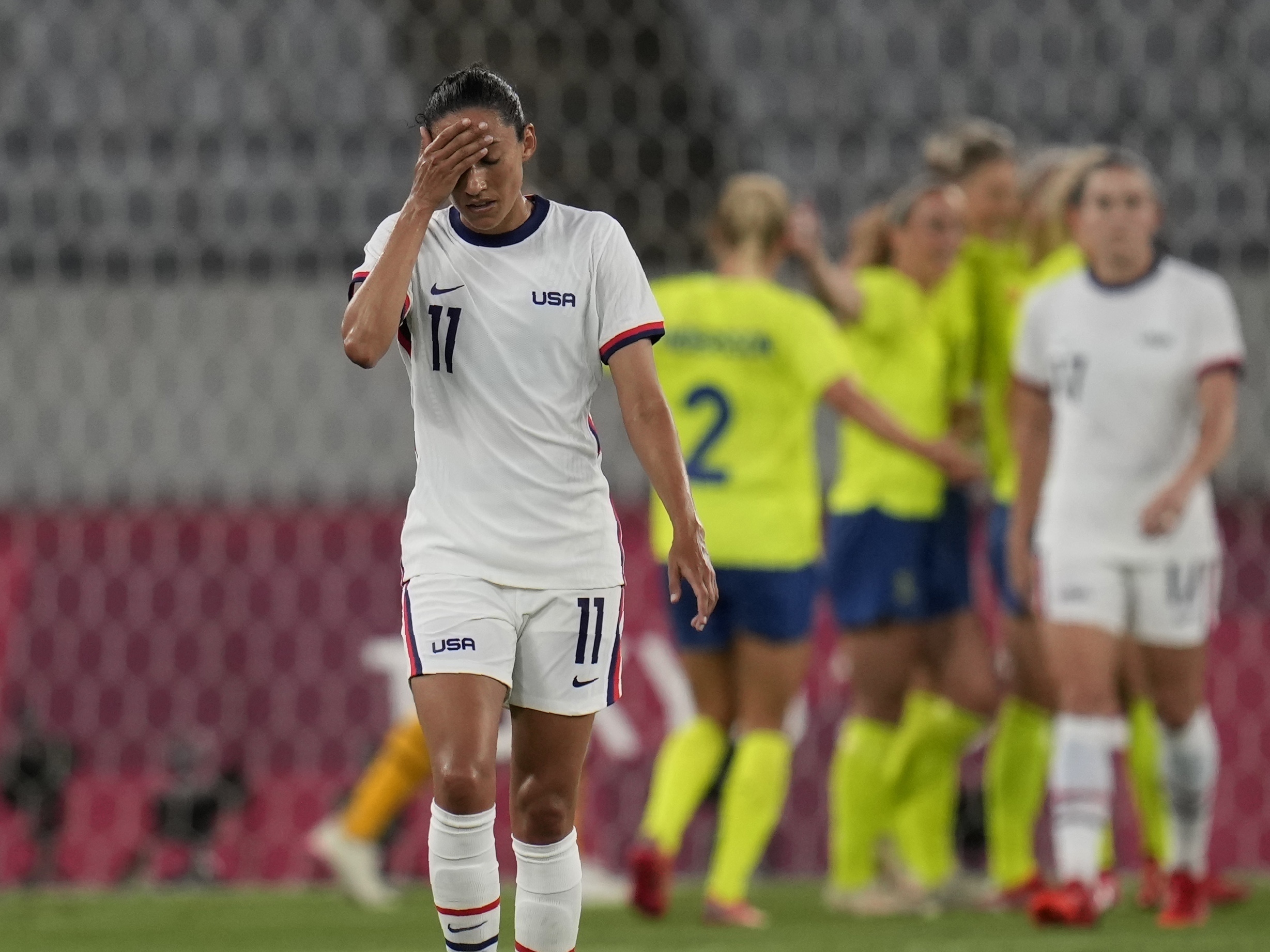 caption: U.S. player Christen Press reacts as Sweden's players celebrate their third goal during a women's soccer match at the Olympics on Wednesday in Tokyo.