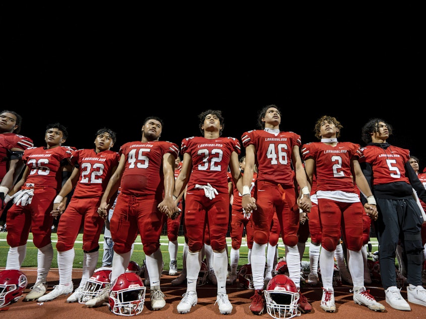caption: FILE - The Lahainaluna High School football team hold hands to thank the fans after a game on Oct. 21, 2023, in Lahaina, Hawaii. Captains of the team whose town was destroyed by a deadly wildfire attended to Super Bowl in Las Vegas as guests of the NFL.