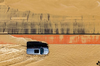 caption: In this aerial picture taken on Aug. 21, a vehicle drives through floodwaters following heavy rains from Tropical Storm Hilary in Thousand Palms, Calif.
