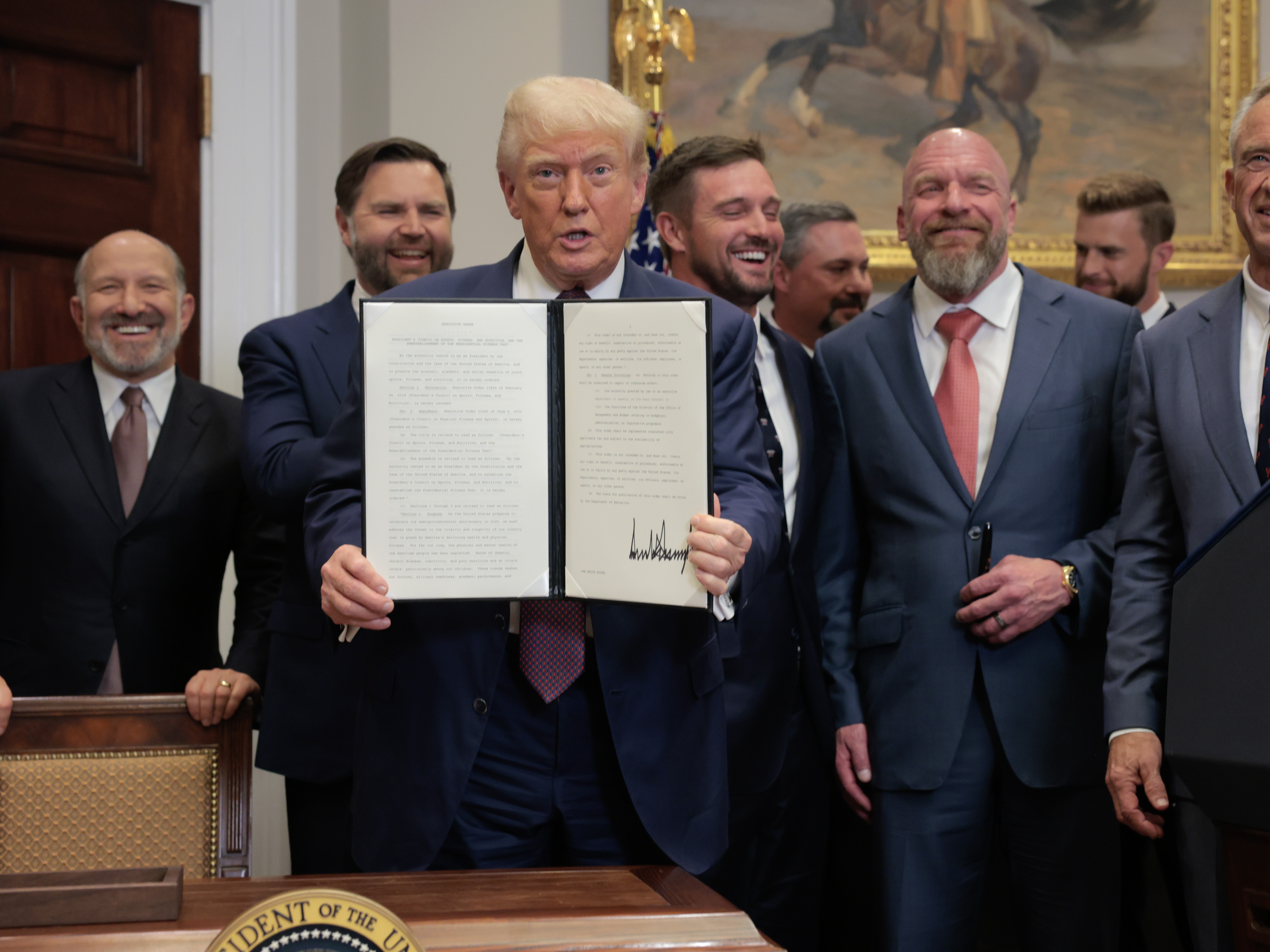 caption: President Trump displays a signed executive order during Thursday's signing ceremony in the White House, surrounded by administration and professional athletes including golfer Bryson DeChambeau and WWE Chief Creative Officer Paul "Triple H" Levesque.