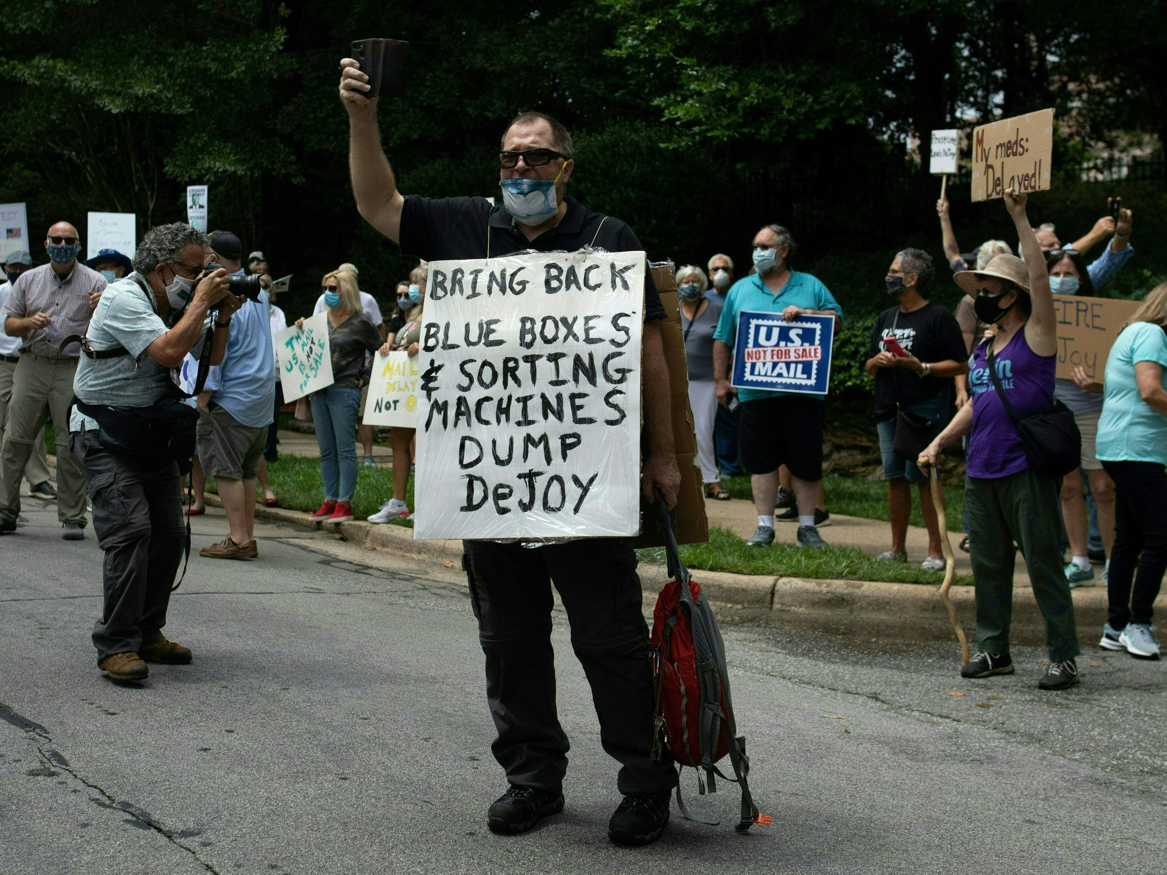caption: A group of protestors holds a demonstration in front of Postmaster General Louis DeJoy's home in Greensboro, N.C. on Sunday. DeJoy has recently come under fire for changes made at the U.S. Postal Service that have delayed mail in advance of the November elections.