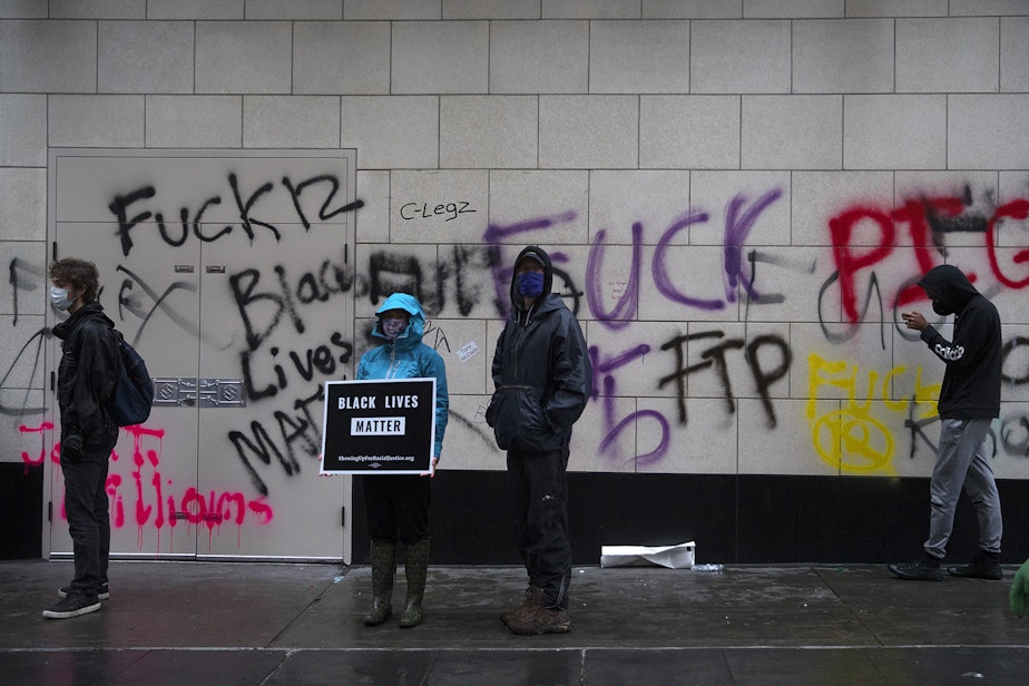 caption: Protesters stand in front of graffiti near the intersection of 5th and Pine Streets on Saturday, May 30, 2020, in downtown Seattle.