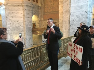 caption: Republican state Rep. Matt Shea records a video on the first day of the legislative session while a small group of supporters gather around him.
