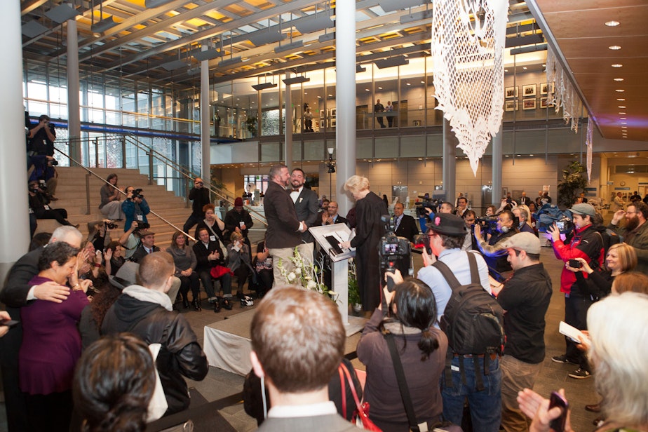 caption: Family, friends and media gather in the Grand Lobby of Seattle’s City Hall to witness over 100 couples take their wedding vows on December 9, 2012.