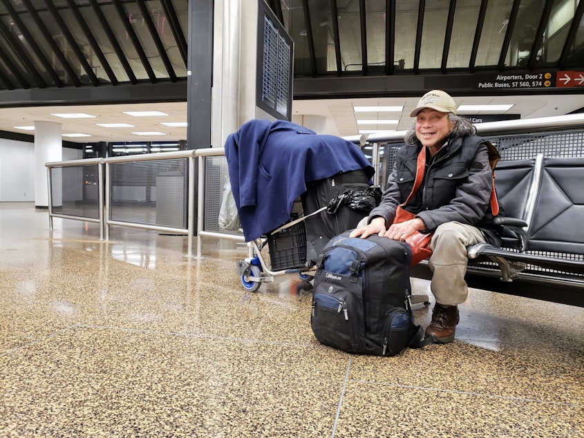 caption: De Chung sits with his bags and belongings near a luggage carousel at SeaTac Airport on Tuesday, January 3, 2023. 