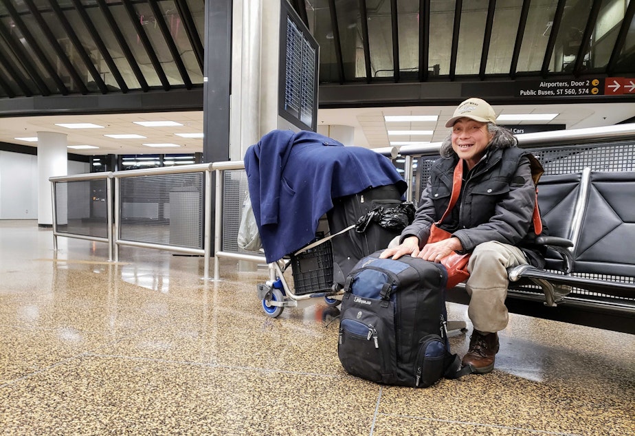 caption: De Chung sits with his bags and belongings near a luggage carousel at SeaTac Airport on Tuesday, January 3, 2023. 