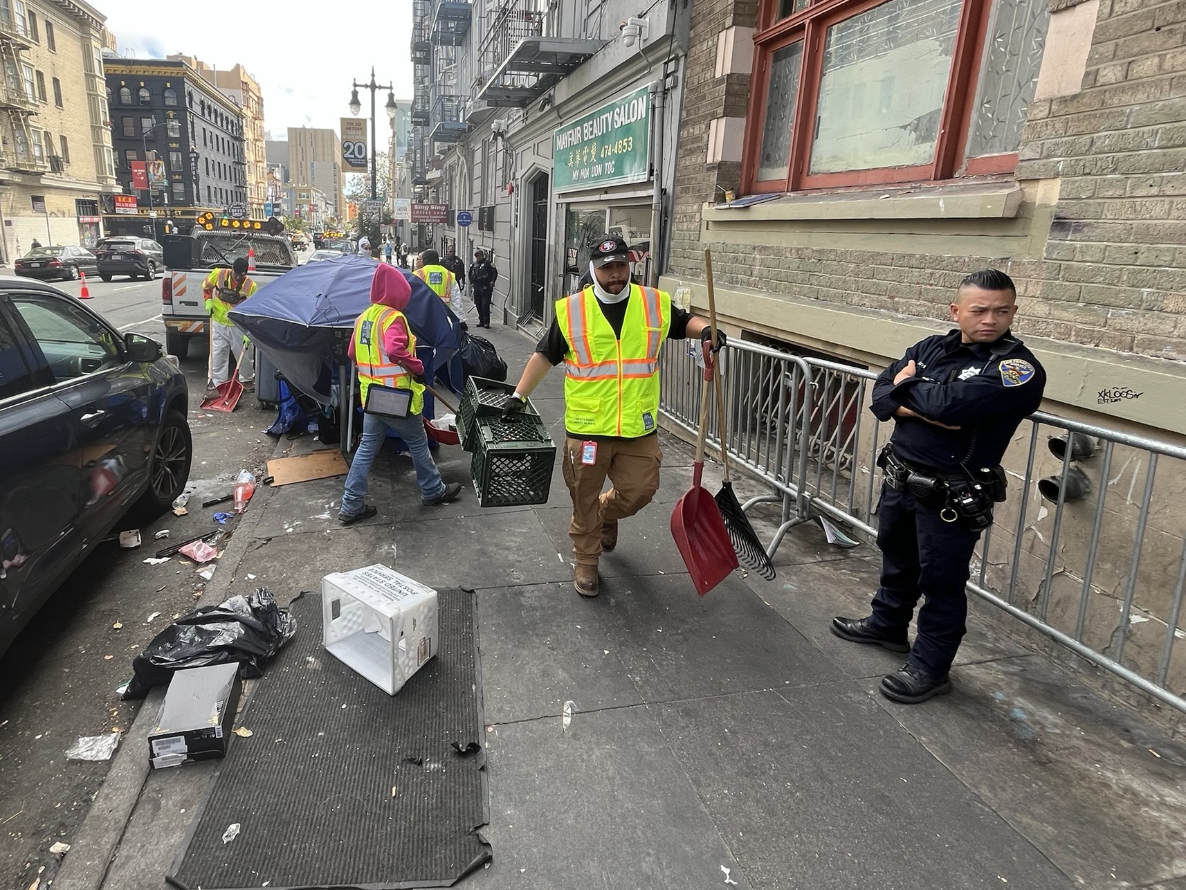 caption: San Francisco police officers keep an eye on city workers as they sweep unsanctioned tents and belongings from a street in the Tenderloin.