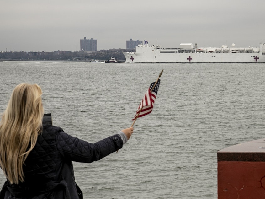 caption: The U.S. Navy hospital ship USNS Comfort is welcomed to New York City by Charlene Nickloan, waving a flag from the Matthew Buono war memorial in Staten Island, N.Y., Monday. Nickloan, who came out with her family, said it was exciting to see the ship, adding "it's so patriotic for us." The ship has some 1,000 beds, and will help New York's health care system during the coronavirus pandemic.