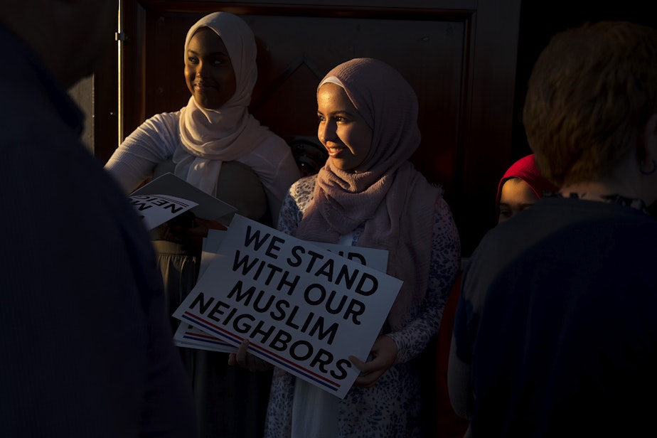 caption: Hibaq Ibrahim, 16, left, and Ihssane El Yacouvi, 14, right, hand out We Stand With Our Muslim Neighbors signs as guests enter the mosque on Monday, March 18, 2019, during an Interfaith Vigil and Anti-Islamophobia Teach-In in response to the mass shooting in Christchurch, New Zealand, at the Muslim Association of Puget Sound in Redmond. 