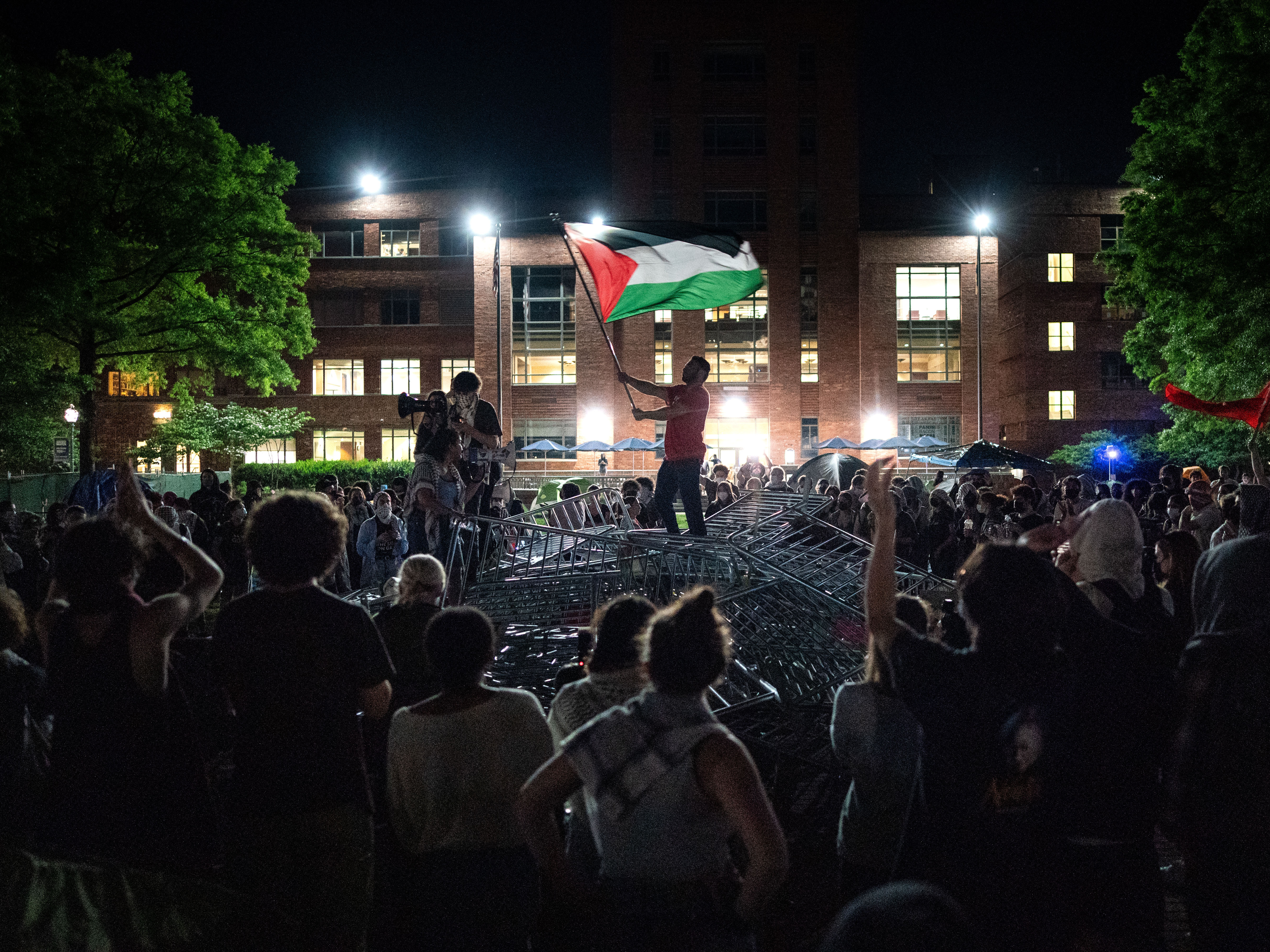 caption: A man holds up a Palestinian flag as activists and students surround piled barricades at an encampment at at George Washington University early Monday.