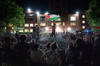 caption: A man holds up a Palestinian flag as activists and students surround piled barricades at an encampment at at George Washington University early Monday.