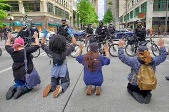 caption: Activists kneel in front Seattle Police during a protest on Sunday, May 31st, 2020.