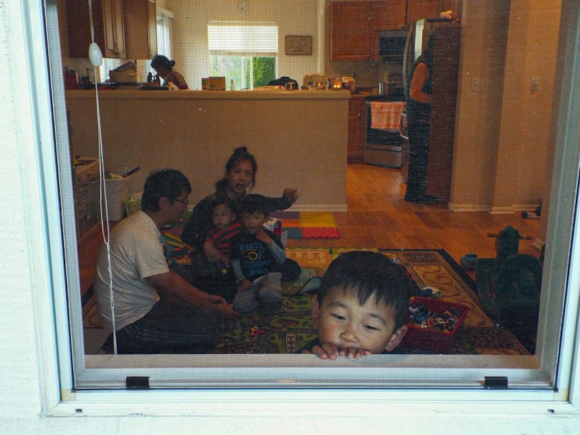 caption: Jonathan Lee, age 3, looks outside his window from his home in Renton, Washington, on June 16, 2020, while his two siblings David and Noel play with their parents in the living room. Behind them, their grandparents wash the dishes and look for a snack in the pantry.