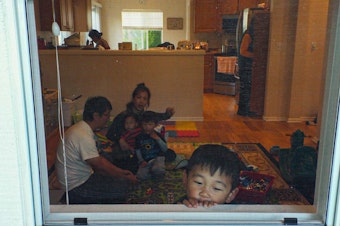 caption: Jonathan Lee, age 3, looks outside his window from his home in Renton, Washington, on June 16, 2020, while his two siblings David and Noel play with their parents in the living room. Behind them, their grandparents wash the dishes and look for a snack in the pantry.