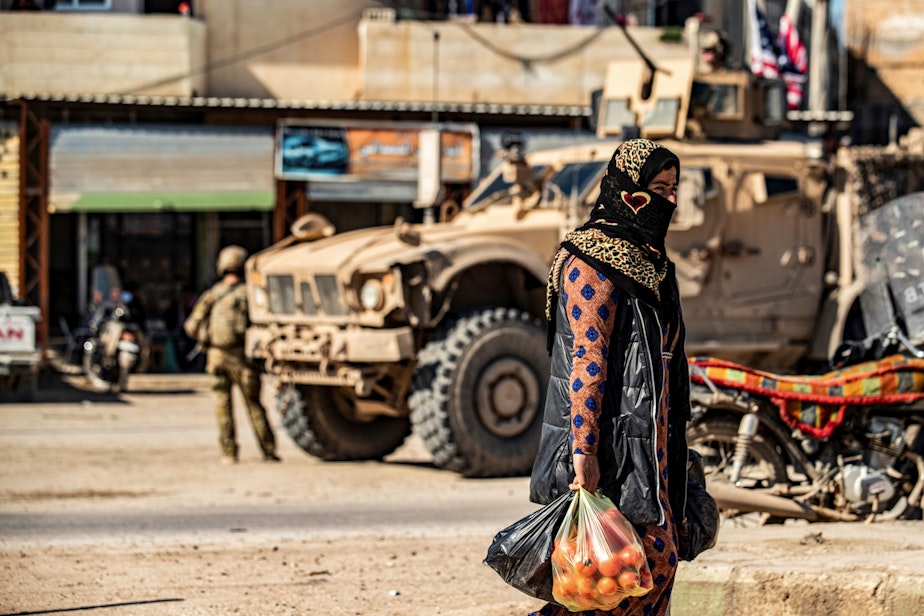 caption: A woman carrying produce walks past a US military armoured vehicle in the town of Tal Tamr along the M4 highway in the northeastern Syrian Hasakeh province, near the border with Turkey, on March 3, 2020. (DELIL SOULEIMAN/AFP via Getty Images)
