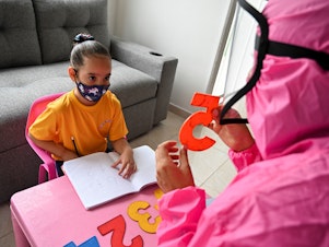 caption: Giving a lesson at the home of a girl in Cali, Colombia, in August 2020, a teacher wears a biosecurity suit to prevent infection from the novel coronavirus. The "teacher at home" program aimed to help students stay in touch with teachers during pandemic school shutdowns.