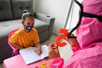 caption: Giving a lesson at the home of a girl in Cali, Colombia, in August 2020, a teacher wears a biosecurity suit to prevent infection from the novel coronavirus. The "teacher at home" program aimed to help students stay in touch with teachers during pandemic school shutdowns.