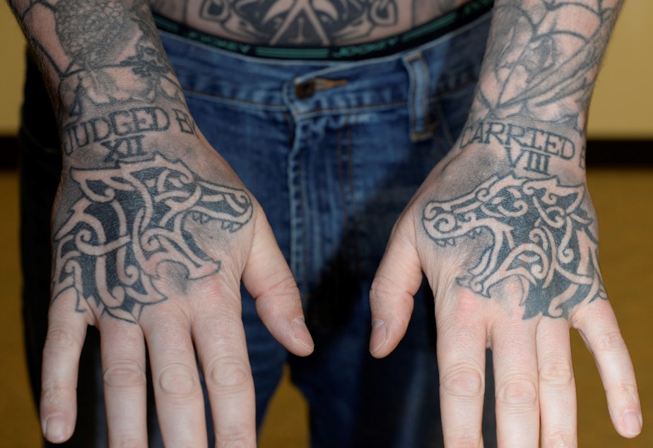 caption: Auburn police officer Jeffrey Nelson displays tattoos on his hands: “JUDGED BY XII” on the right hand. “CARRIED BY VIII” on the left. That’s shorthand for an old policing proverb: “I’d rather be judged by 12” jurors than “carried by six” — or sometimes eight — pallbearers.