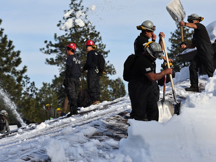 caption: Members of the California Army National Guard Joint Task Force Rattlesnake shovel snow from a rooftop after a series of winter storms dropped more than 100 inches of snow in the San Bernardino Mountains in Southern California on March 8, 2023 in Crestline, California.