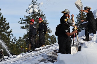 caption: Members of the California Army National Guard Joint Task Force Rattlesnake shovel snow from a rooftop after a series of winter storms dropped more than 100 inches of snow in the San Bernardino Mountains in Southern California on March 8, 2023 in Crestline, California.