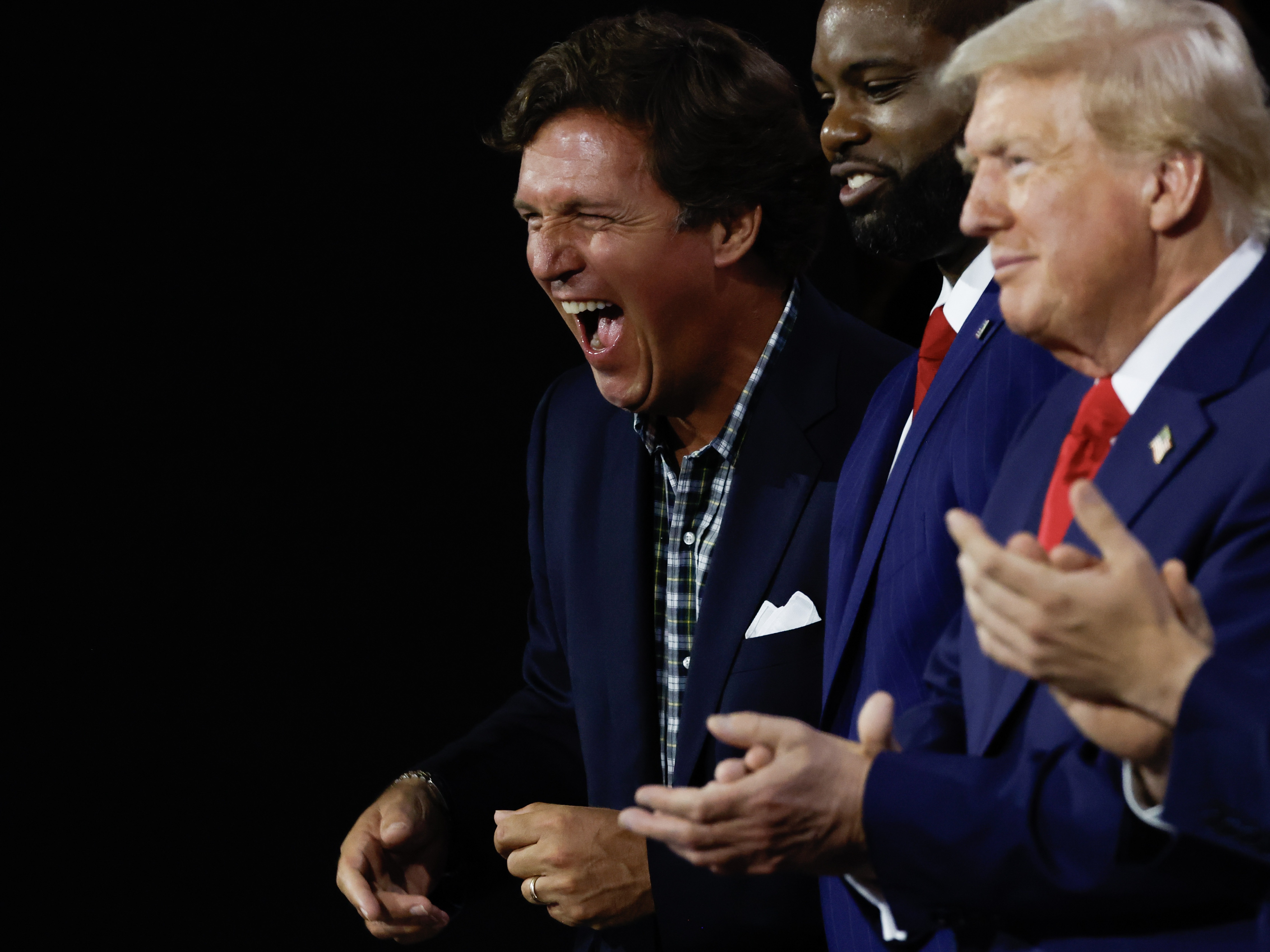 caption: Right-wing personality Tucker Carlson laughs as he stands alongside Congressman Byron Donalds, Republican presidential candidate Donald Trump, and vice presidential candidate J.D. Vance on the first day of the Republican National Convention in Milwaukee.
