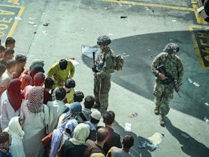 caption: U.S. soldiers stand guard as Afghan people wait at the Kabul airport in Kabul on Aug. 16, 2021, after a stunningly swift end to Afghanistan's 20-year war, as thousands of people mobbed the city's airport trying to flee the group's feared hardline brand of Islamist rule.