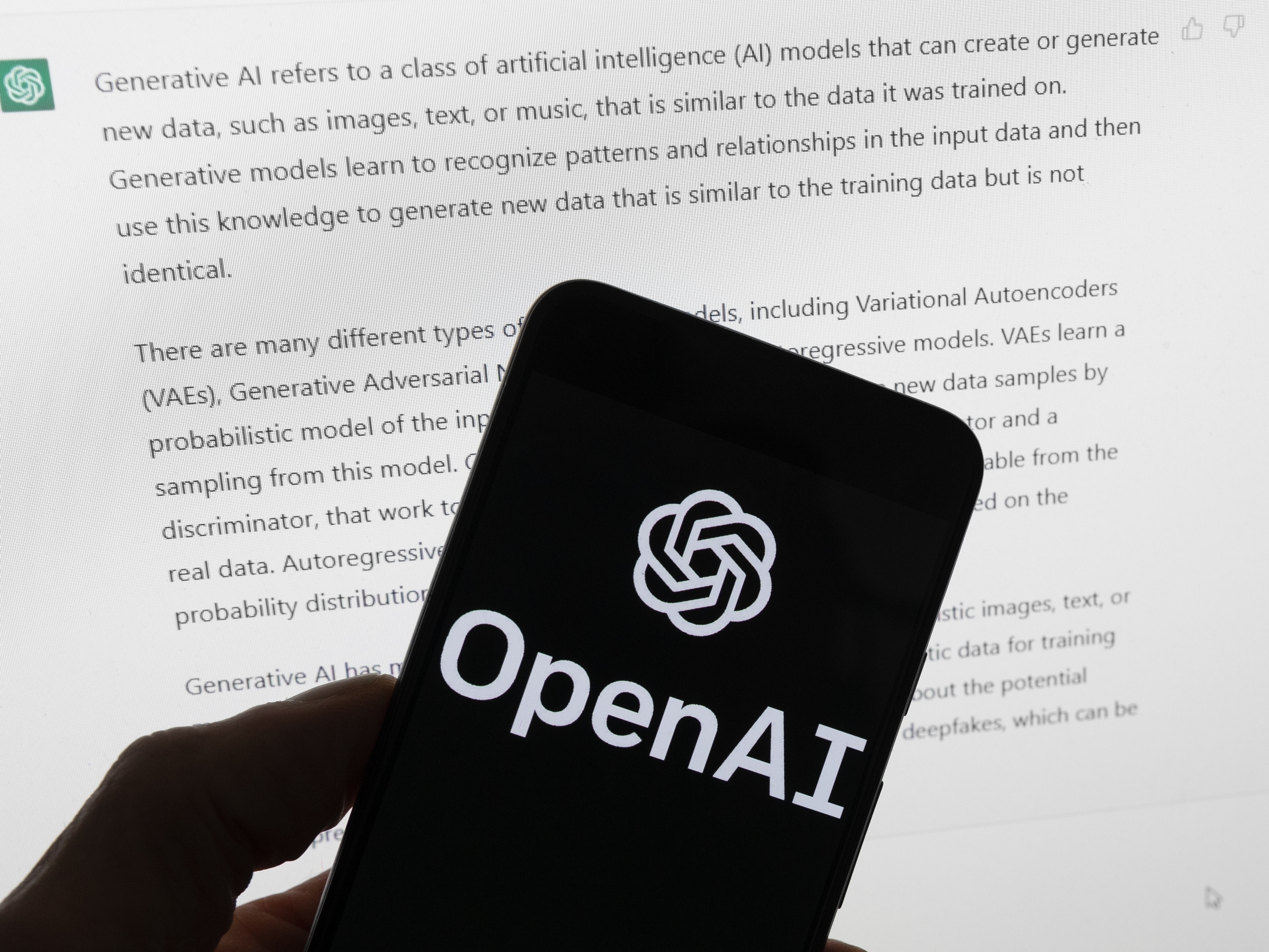 caption: OpenAI's logo is on the screen of a mobile phone that's being held in front of a computer screen displaying output from ChatGPT.