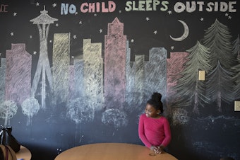caption: Mariah Hicks, 7, stands in front of a chalk drawing depicting downtown Seattle in a common area at Mary's Place, a homeless shelter.