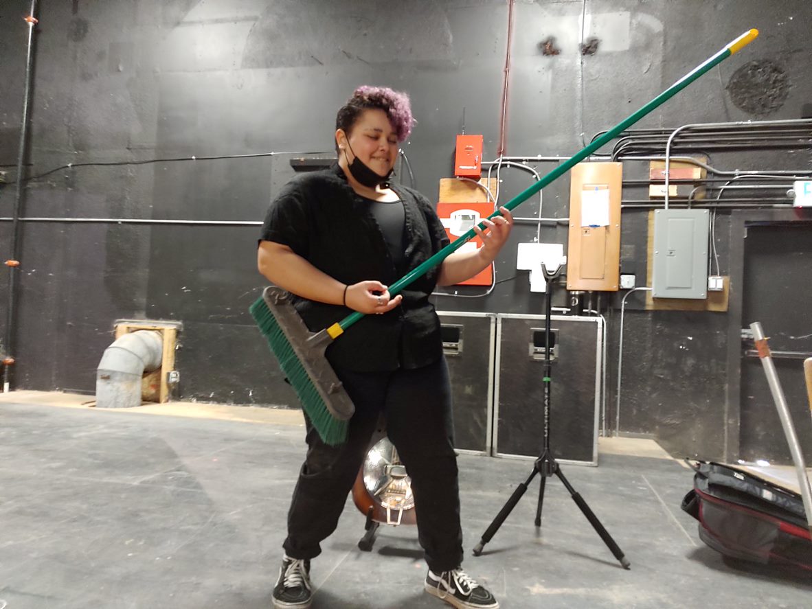 caption: Ari Whidbey playing a broom like a guitar onstage before a Rhapsody Project event at the Black and Tan Hall in Seattle, on March 5th, 2023.