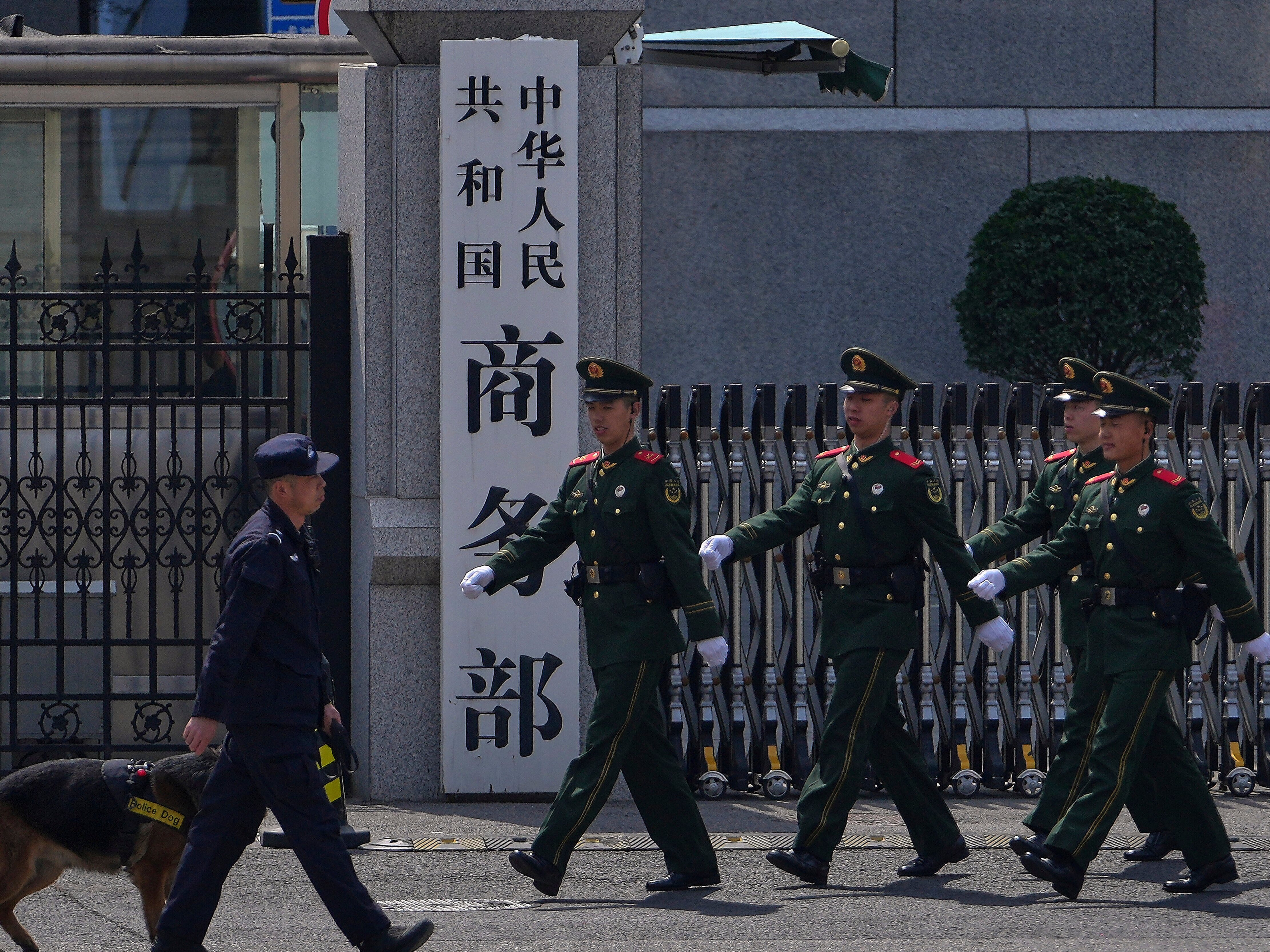 caption: FILE - Paramilitary soldiers and a police officer with a sniffer dog march past the main entrance gate of China's Ministry of Commerce, in Beijing, on April 3, 2025.