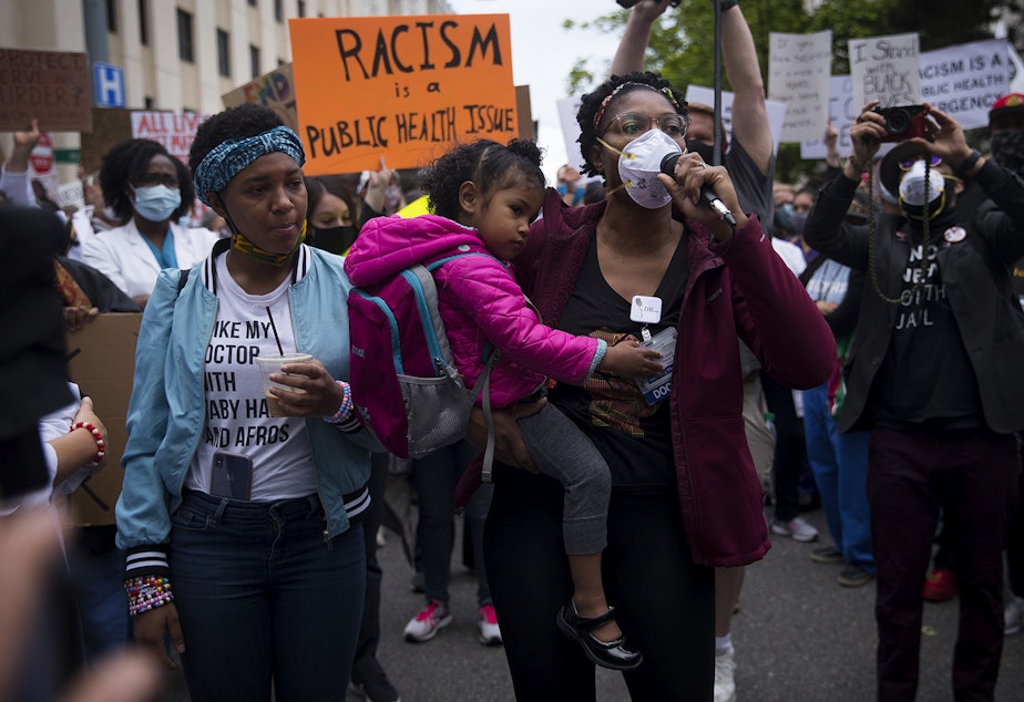 caption: Organizer Dr. Estell Williams, center, holds daughter Estella while speaking to a crowd of thousands who gathered in protest of police violence and to demand justice before marching from Harborview Medical Center to Seattle City Hall on Saturday, June 6, 2020, in Seattle. 