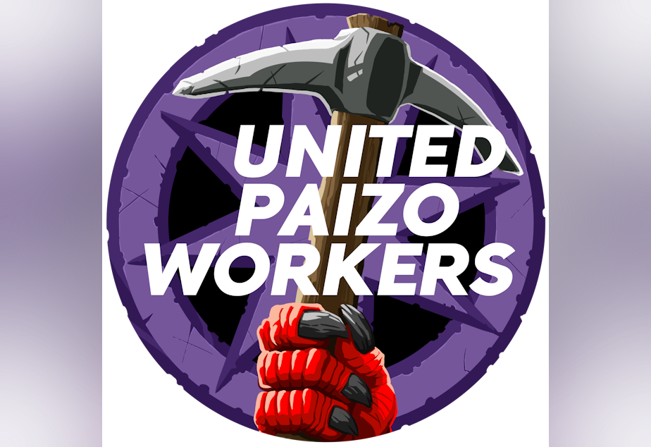 caption: The logo for United Paizo Workers, which includes a red, clawed hand holding a pickaxe in front of a purple starred background.
