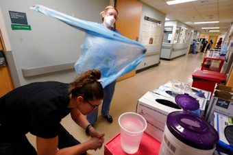 caption: Registered nurse Jessalynn Dest, left, removes protective equipment and washes her hands after leaving a Covid-19 patient's room as speech therapist Sam Gibbs puts on safety clothing while preparing to see a patient in the acute care unit of Harborview Medical Center, Friday, Jan. 14, 2022, in Seattle.