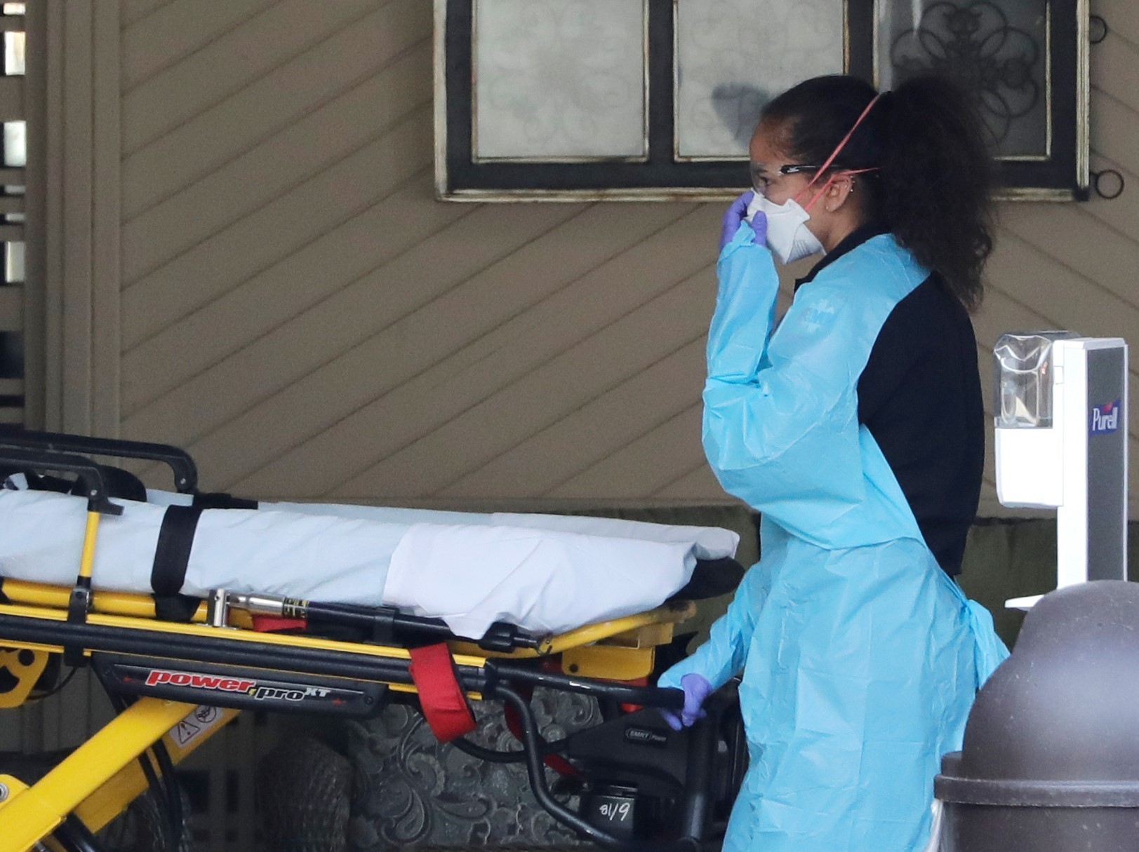 caption: An ambulance worker adjusts her protective mask as she wheels a stretcher into a nursing facility in Kirkland, Wash., on Saturday where more than 50 people were found to be sick and are being tested for COVID-19 virus.