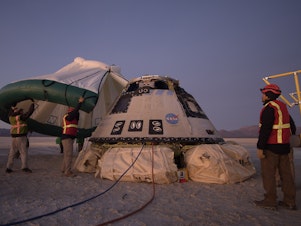 caption: Boeing, NASA, and U.S. Army personnel work around the Boeing Starliner spacecraft shortly after it landed in White Sands, N.M., Sunday.