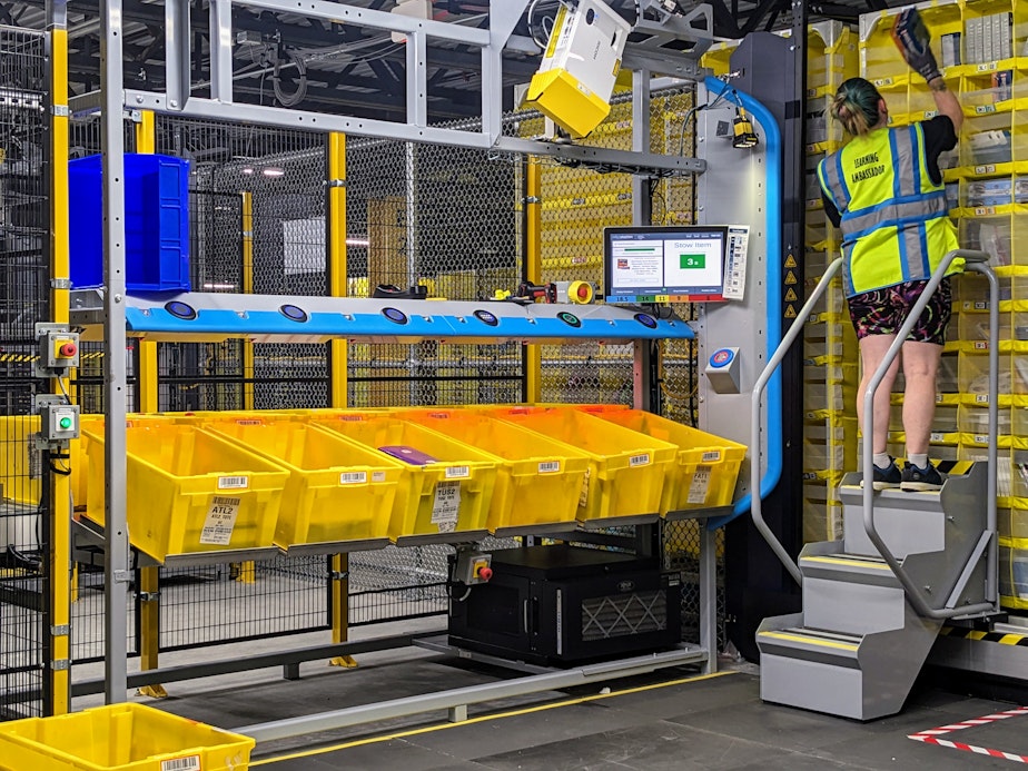 caption: An Amazon associate uses a latter to stow products on a high shelf at PAE2, a robotics facility on Arlington, Wash.