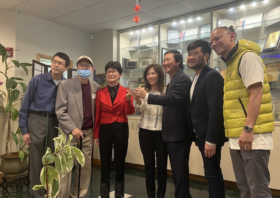 caption: Northwest Asian Weekly publisher Assunta Ng (third from left) passes exchanges a key representing leadership behind the news outlet to the new publisher, Grace Roh (center.) From left to right: John Liu, George Liu, Assunta Ng, Grace Roh, Jeffrey Roh, Sam Cho, and Tim Wang. 