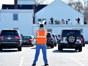 caption: Volunteer Al Wheeler, wearing bunny ears, keeps an eye on the service and cars during a drive-in Easter service at First Baptist Church in Plaistow, N.H., on April 12. Since the COVID-19 pandemic has made services inside the church a danger to health, the church started holding gatherings in its parking lot.