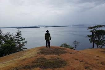 caption: KUOW's John Ryan looks out at Canada's Moresby and Saltspring islands from Turn Point, Stuart Island, Washington, in 2019. Washington's deepest spot is hidden beneath the waves in between.