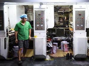 caption: Workers in green polo shirts and blue caps monitor machines making plastic products at the Dongguan Fangjie Printing and Packaging Company.