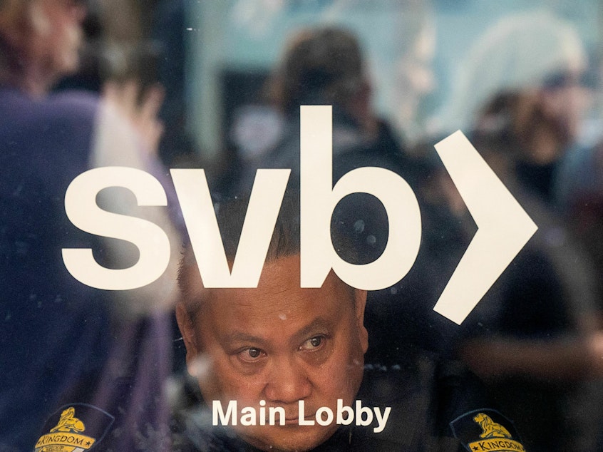 caption: A security guard looks out a door as customers line up at Silicon Valley Bank headquarters in Santa Clara, California on March 13, 2023.