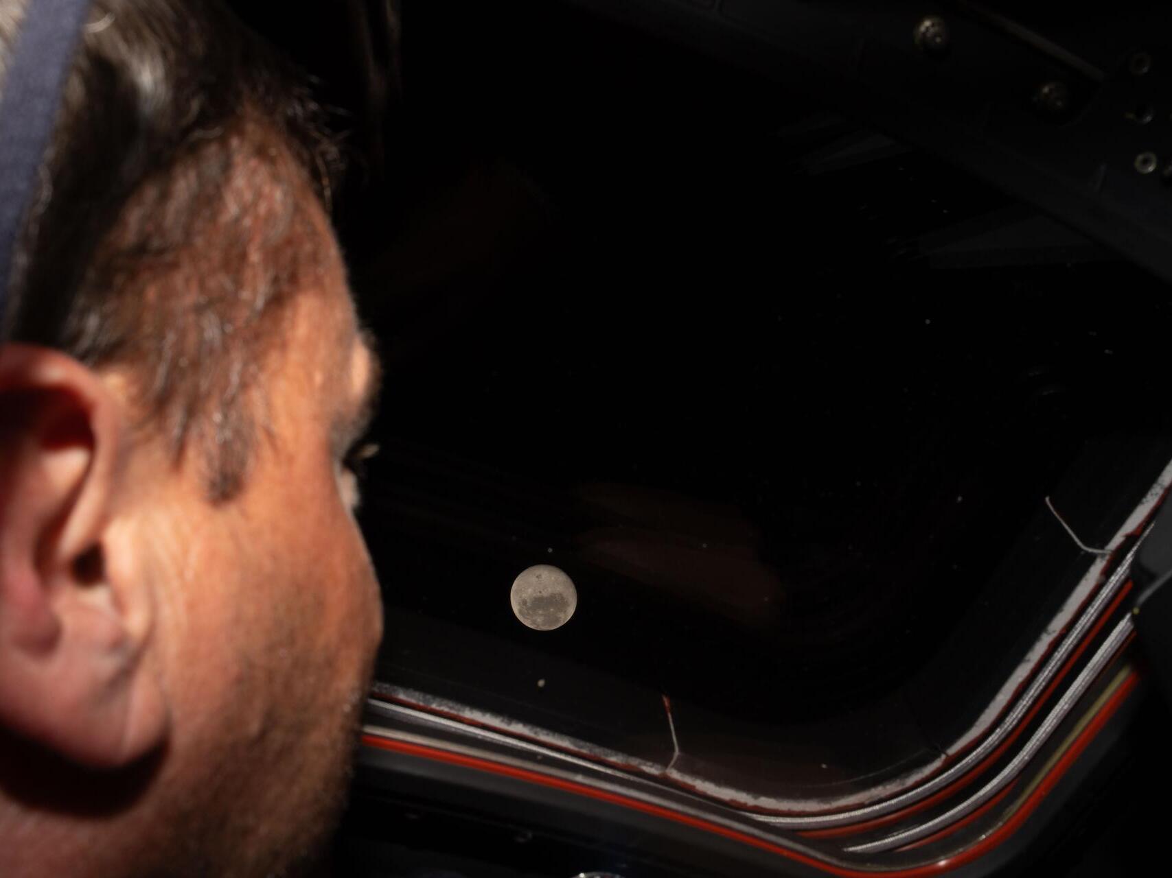 caption: Artemis II commander and NASA astronaut Reid Wiseman looks out one of the Orion spacecraft's main cabin windows at the moon ahead of the crew's lunar flyby. After successfully looping around the moon, the space capsule is now headed back toward Earth.