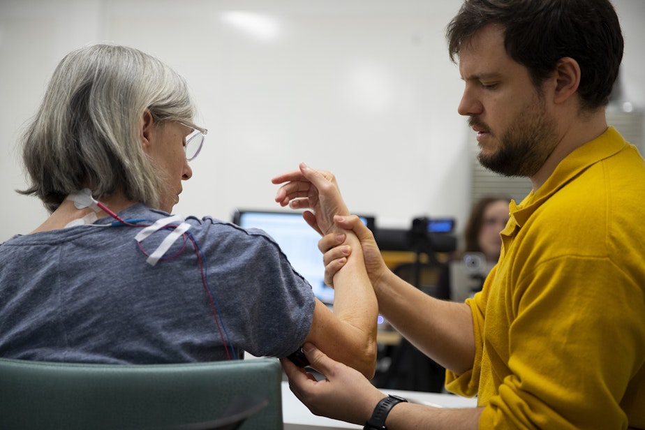 caption: Research assistant Adrià Robert-Gonzalez, right, adjusts participant Debbie Strom's arm after placing electrodes on the back of her neck before a non-invasive transcutaneous spinal cord simulation session, on Monday, April 15, 2024, at Wallace Hall on the University of Washington campus in Seattle. 