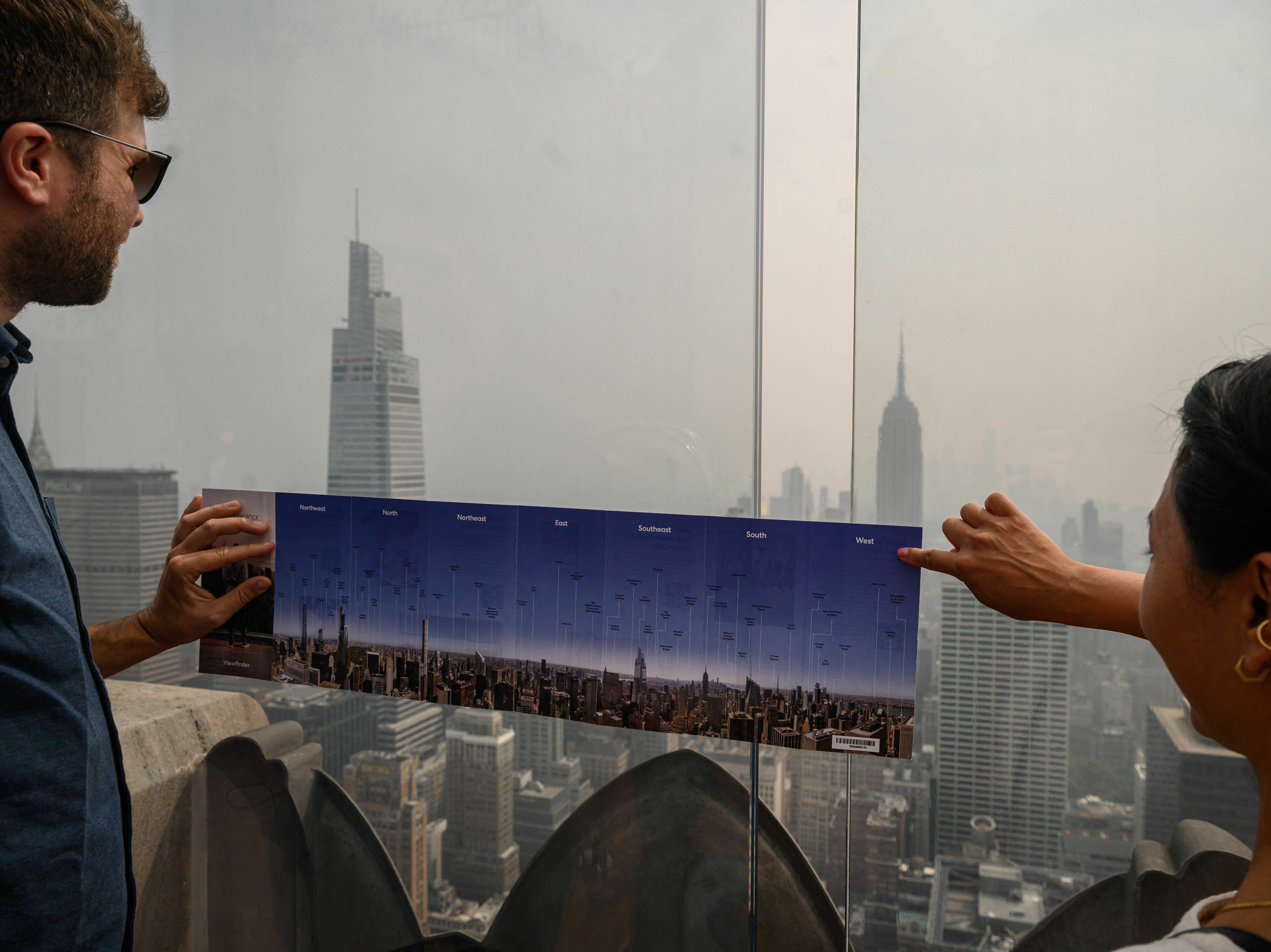 caption: Heavy smoke from Canada's wildfires has put a thick haze over large parts of the U.S. this summer. Here, visitors to New York City hold a map showing city landmarks on a clear day as they stand on the viewing deck of Rockefeller Center in late June.