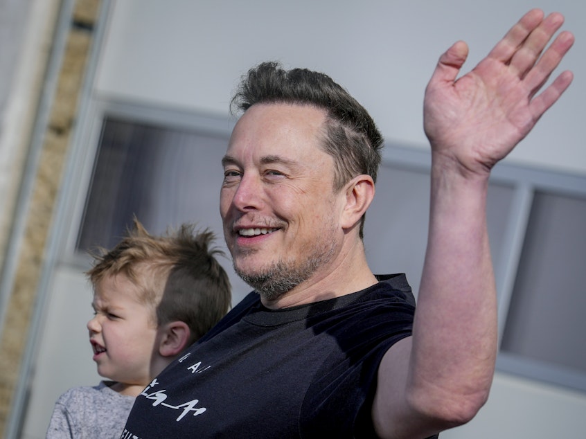 caption: With his son in one arm, Tesla CEO Elon Musk waves while visiting the Tesla Gigafactory in Germany in March.