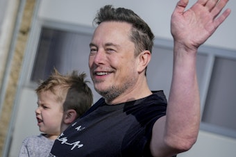caption: With his son in one arm, Tesla CEO Elon Musk waves while visiting the Tesla Gigafactory in Germany in March.